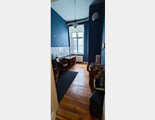 Schmaler, länglicher Raum mit Holzdielenboden und dunkelblau gestrichenen Wänden. Der Raum enthält einen kleinen Holzschreibtisch, zwei braune Stühle, eine Hängelampe, weiße Wandfliesen in der Mitte der Wände sowie ein großes Fenster, das Tageslicht hereinlässt.