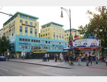 Gelbes, mehrstöckiges Gebäude mit blauen Fassadenelementen und Beschriftung „Ärzte Schönhauser“ im urbanen Umfeld, daneben ein kleiner Imbissstand mit buntem Dach und Sitzgelegenheiten; im Vordergrund Menschen auf einem Gehweg sowie eine Straße mit parkenden Autos.