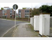 Straßenszene in einem Wohngebiet mit modernen Mehrfamilienhäusern aus rotem Backstein mit großen Fenstern im Hintergrund. Im Vordergrund ein Gehweg mit weißem Zaun und Mauern, daneben eine Kurve mit Straßenschildern.