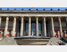 Fassade des Alten Museums in Berlin mit hohen korinthischen Säulen, einem lateinischen Schriftzug im Architrav und der deutschen Flagge auf dem Dach. Im Vordergrund steht ein Brunnen aus Stein, während Treppenstufen zum Eingang des Gebäudes führen.
