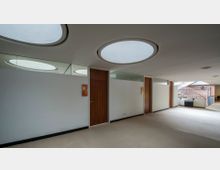 Ein moderner Innenraum mit hellen Wänden, runden Oberlichtern in der Decke, Holztüren und Glaswänden. Im Hintergrund ist eine Treppe mit Geländer zu sehen, die zu einer höheren Ebene führt.