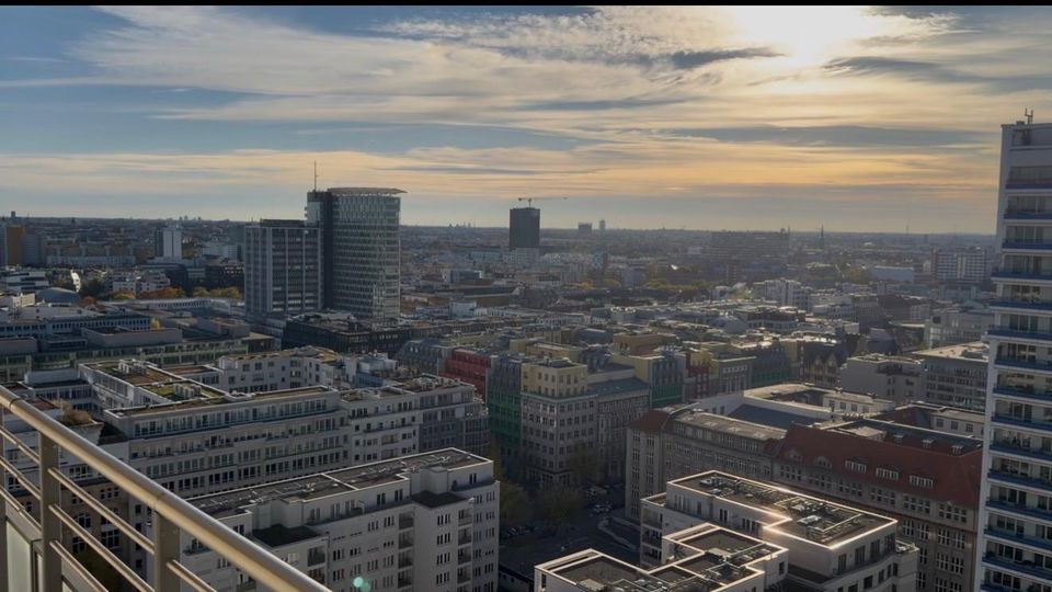 Luftaufnahme einer städtischen Landschaft mit zahlreichen Gebäuden unterschiedlicher Höhe, darunter moderne Hochhäuser und farbige Altbauten. Im Hintergrund ein weitläufiger Blick über die Stadt mit einem klaren Himmel und vereinzelten Wolken bei Sonnenuntergang.