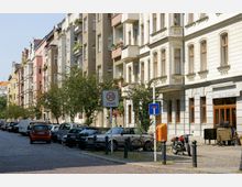 Straße mit mehrstöckigen Wohngebäuden im Altbaustil auf beiden Seiten, pastellfarben gestrichen und mit Balkonen versehen. Am Straßenrand parken Autos, ein "30-Zone"-Schild ist sichtbar, Bäume säumen die gepflasterte Straße, und ein gelber Postbriefkasten sowie ein Verkehrsschild für eine Sackgasse befinden sich im Vordergrund.