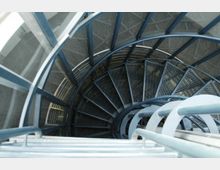 Metallische Wendeltreppe mit Gitterstufen, die in die Tiefe führt, umgeben von einer Glaswand und blauen Metallgeländern. Der Boden und Wände im Hintergrund bestehen aus grauem Material.