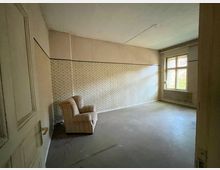 Leerer Innenraum eines unsanierten Zimmers mit grauem Boden, gemusterter Tapete und hohen Decken. Rechts befindet sich ein großes Sprossenfenster über einem Heizkörper; links ist eine Tür und ein einzelner Sessel steht in der Ecke.