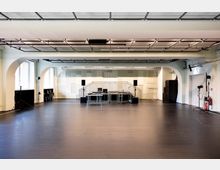 Großer, leerer Studio- oder Proberaum mit dunklem Boden, weiß gestrichenen Wänden und breiten Rundbögen. Im Hintergrund steht eine kleine Bühne mit Tischen, Monitoren und Lautsprechern; an der Decke sind Schienen mit Beleuchtung und Technik montiert, seitlich befinden sich Fenster und Equipment.