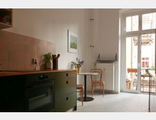 Helle Küche mit weißem Fliesenboden und pastellfarbenem Fliesenspiegel an der Wand. Links eine grüne Küchenzeile mit Holzarbeitsplatte, rechts ein runder Tisch mit zwei Holzstühlen vor bodentiefen Fenstern, die auf einen Balkon führen.