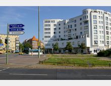 Straßenszene mit modernem weißen Wohn- und Geschäftsgebäude im Vordergrund, flankiert von niedrigeren Gebäuden im traditionellen Stil mit roten Dächern. Wegweiser zeigen die Richtungen zu Tempelhof, Steglitz, Dresden und Schönefeld, während im Hintergrund eine U-Bahn-Station („U-Bahn Innsbrucker Platz“) sichtbar ist.