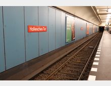 U-Bahn-Station mit gleisnahem Bahnsteig und blauer Wandverkleidung, an der mehrere Werbeplakate sowie ein rotes Schild mit der Aufschrift „Hallesches Tor“ angebracht sind. Rechts im Bild ist ein beleuchteter Bereich mit Sitzgelegenheiten sichtbar.