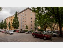 Mehrstöckige Wohngebäude mit Putzfassaden und Balkonen, umgeben von Bäumen. Im Vordergrund eine gepflasterte Straße mit parkenden Autos und Fahrrädern sowie einer alten Straßenlaterne.