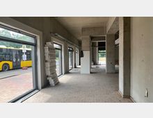 Leerer, unfertiger Innenraum mit Betonwänden, großen Glasfenstern und Fliesenboden; durch die Fenster ist ein gelber Linienbus sowie eine Bushaltestelle zu sehen.