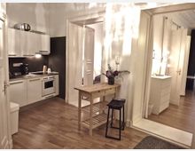 Wohnraum mit integrierter Küche, ausgestattet mit weißen Hängeschränken, einem Herd und einer Spüle. Der Raum verfügt über einen Holzboden, eine Holzkonsole mit Dekoration, sowie eine offene Verbindung zu einem weiteren Raum mit einer Kommode und Spiegel.