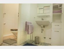 Kleines Badezimmer mit weiß gefliesten Wänden und einem Waschbecken mit Wandspiegel. Links befindet sich eine separate Toilettenecke, daneben ein Handtuchständer mit lila Handtüchern, ein Mülleimer aus Metall und ein weißer Unterschrank. Der Boden ist mit gemusterten Fliesen in verschiedenen Farben gestaltet.