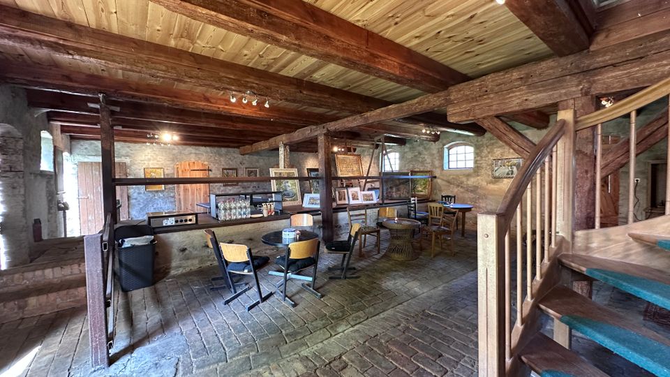 Innenraum eines rustikalen Cafés in einem umgebauten Stein- und Fachwerkgebäude mit sichtbaren Holzbalken, Kopfsteinpflasterboden und kleinen Tischen mit Stühlen. Im Raum stehen eine Theke mit Flaschen, gerahmte Bilder an den Steinwänden sowie eine Holztreppe mit Geländer im Vordergrund.