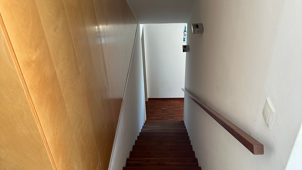 Blick in ein schmales, innenliegendes Treppenhaus mit dunklen Holzstufen, weiß gestrichenen Wänden und Handlauf rechts. Links verläuft eine große Holzverkleidung, am Ende führt die Treppe zu einem kleinen Flur mit Holzfußboden.