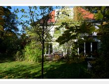 Zweistöckiges Wohnhaus mit rotem Dach, umgeben von einem üppigen Garten mit Rasen, Bäumen und Sträuchern. Im Vordergrund sind Blätter und Äste sichtbar, die teilweise den Blick auf das Haus verdecken.