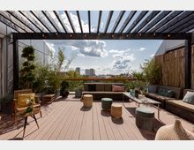 Moderne Dachterrasse mit Holzfußboden, einer schwarzen Pergola mit Lichterkette, Sitzmöbeln und dekorativen Pflanzen. Im Hintergrund ist eine Stadtlandschaft mit Gebäuden und bewölktem Himmel zu sehen.