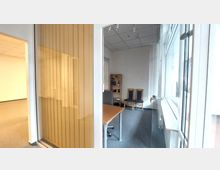 Blick in ein Büro mit Glastür und vertikalen Lamellen, einem Schreibtisch mit Bürostuhl, zwei Stühlen mit gepolsterter Sitzfläche und einem Regal an der Rückwand. Der Raum hat helle Wände, einen Teppichboden und ein großes Fenster auf der rechten Seite.