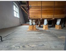 Blick in den Dachboden einer historischen Mühle mit Holzdielenboden, einer Holzkonstruktion für Getreideverarbeitung und Rohrleitungen, die in Holzbehälter mit Trichtern führen. Links befindet sich ein Fenster mit gewölbtem Rahmen, durch das Tageslicht einfällt.