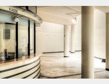 Schalterhalle mit modernem Kassenbereich aus Glas und Metall, daneben eine weite, helle Fläche mit Säulen und cremefarbenen Wänden. An der Wand sind Schilder mit Nummerierungen erkennbar.