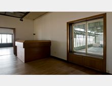 Ein kleiner Raum mit Holzboden, einer Holztheke und einer großen Fensterscheibe, die in eine industrielle Halle mit hohen Decken und großen Fenstern blickt. Rechts unterhalb des Fensters befinden sich Holzeinbauten, und links führt eine Tür in einen weiteren Raum.