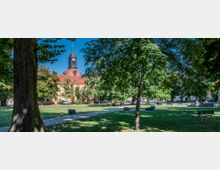 Parklandschaft mit großen Bäumen, Rasenflächen, Bänken und einem gepflasterten Weg. Im Hintergrund ist ein historisches Gebäude mit rotem Ziegeldach und einem Turm mit Spitzdach zu sehen.