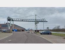 Industriegebiet mit einem Hafenkran und einem langgestreckten Backsteingebäude im Hintergrund. Im Vordergrund verläuft eine asphaltierte Straße, daneben geparkte Fahrzeuge sowie eine Grünfläche am Wasserufer.