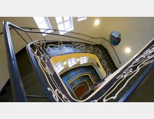 Treppenhaus mit geschwungenem Geländer aus dekorativem Metall, das weiß bemalt ist, und in die Tiefe führenden Stufen. Die Wände sind gelblich gestrichen, mit runden Leuchten versehen, und oben sind Fenster mit Tageslichteinfall sichtbar.