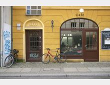 Fassade eines gelben Gebäudes mit einer braunen Holztür links und einem Caféeingang mit großen Glasfenstern rechts. Davor stehen zwei Fahrräder, und an der Wand ist ein Graffiti sowie eine Schiefertafel mit einer Menüaufschrift angebracht.