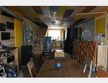 Ein multifunktionaler Innenraum mit bunter Holzdecke und einer rustikalen Holztreppe, die zu einem Fenster mit Pflanzen führt. Diverse Einrichtungsgegenstände wie ein Holzofen, Regale mit Dosen und künstlerische Dekorationen prägen den Raum, während ein großer Holztisch im Vordergrund steht.