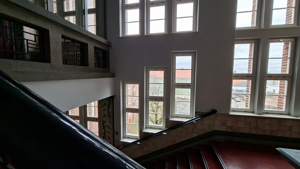 Helles Treppenhaus in einem mehrstöckigen Gebäude mit großen, rechteckigen Fensterfronten und Blick auf umliegende Backsteinbauten. Zu sehen sind ein Treppenlauf mit dunklem Geländer, geflieste Wandbereiche und ein offener Zwischenbereich mit Oberlichtfenstern.