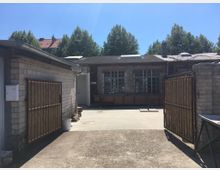 Ein Hof mit gepflastertem Boden, umgeben von niedrigen Backsteingebäuden mit großen, vergitterten Fenstern. Im Vordergrund befindet sich ein geöffnetes Tor aus Holz und Metall, im Hintergrund stehen Bäume und Dächer von Wohnhäusern.