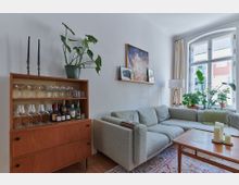 Wohnzimmer mit einer grauen Eck-Couch, einem Holztisch und einem Teppich mit dekorativem Muster. Links steht ein Holzregal mit Gläsern und Spirituosen, während auf der Fensterbank und in der Ecke mehrere Zimmerpflanzen platziert sind. Ein großes Fenster mit weißen Rahmen lässt viel Tageslicht in den Raum.
