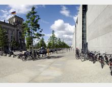 Straßenszene in Berlin mit dem Reichstagsgebäude links im Bild, gekennzeichnet durch die deutsche Flagge auf dem Dach, und einer modernen Betonfassade rechts. Im Vordergrund sind Fahrradständer, parkende Autos und eine gepflasterte Promenade mit kleinen Bäumen zu sehen.