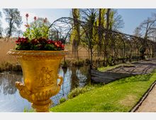 Goldene Blumenvase mit roten Blumen im Vordergrund, stehend an einem kleinen Gewässer in einem Park. Im Hintergrund befindet sich ein Rankgerüst aus Metall, umgeben von Bäumen und Rasenflächen.