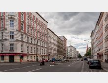 Straßenszene in einem städtischen Wohngebiet mit mehrstöckigen Altbauten im klassischen Stil, überwiegend in Weiß und Rot gehalten. Auf der Straße befinden sich geparkte Autos, ein Motorradfahrer, sowie einzelne Bäume und Straßenlampen; im Hintergrund sind Wolken und ein Kirchturm zu sehen.