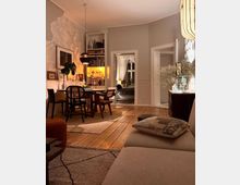 Gemütliches Wohnzimmer mit Holzboden und skandinavischem Einrichtungsstil. Im Vordergrund ein helles Sofa, im Hintergrund ein Esstisch mit Stühlen, eine beleuchtete Bücherwand und eine Tür, die in ein weiteres Zimmer führt.