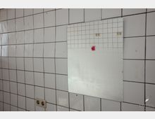 Wand mit weißen, glänzenden Fliesen in einem vermutlich industriellen oder sanitären Raum; ein rechteckiger, einfacher Spiegel ist an der Wand angebracht, daneben eine Steckdose und Spuren von Abnutzung.