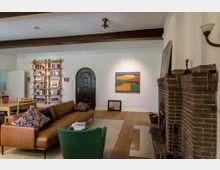 Ein stilvoll eingerichtetes Wohnzimmer mit Holzbalkendecke, einer braunen Ledercouch und grünem Sessel im Vordergrund. An der rechten Wand ein gemauerter Kamin aus Ziegelsteinen, links eine Bücherwand aus Holzregalen, zentral ein Gemälde in warmen Farben und eine dunkle, halbrunde Tür.