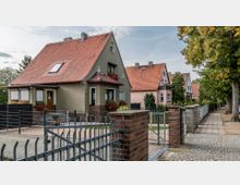 Eine Reihen von Einfamilienhäusern mit roten Ziegeldächern und Vorgärten, die von Metallzäunen und Klinkermauern gesäumt sind. Im Vordergrund führt ein gepflasterter Gehweg entlang der Grundstücke, flankiert von großen Bäumen.