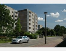 Mehrstöckiges Wohngebäude aus Plattenbau mit sichtbaren Balkonen, umgeben von Bäumen und Grünflächen. Im Vordergrund eine Straße mit einem geparkten silbernen Auto und einer Straßenlaterne, unter einem blauen Himmel mit vereinzelten Wolken.