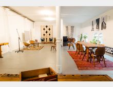 Großer, heller Raum mit modernem und vintage-inspiriertem Interieur, ausgestattet mit Holzstühlen, Tischen und verschiedenen Teppichen. An den weißen Wänden hängen Schwarz-Weiß-Bilder und dekorative Wandteppiche, während große Fenster und Grünpflanzen das Ambiente beleben.