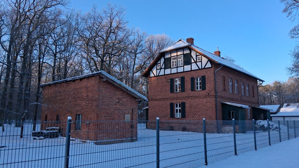 Zwei Gebäude aus rotem Backstein, eines mit Fachwerkelementen, stehen in einer verschneiten Landschaft, umgeben von einem Metallzaun und kahlen Bäumen. Der Himmel ist klar und blau.