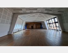 Großer, leerer Veranstaltungssaal mit Holzboden und hoher, schallgedämmter Decke mit eingelassenen Leuchten. Rechts sorgt eine hohe Fensterfront für Tageslicht, hinten ist eine holzverkleidete Stirnwand mit Tür und seitlichen Vorhängen zu sehen.