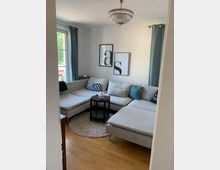 Wohnzimmer mit einer großen, hellgrauen Eckcouch, runden Beistelltisch in dunkler Holzoptik und beigefarbenem Teppich auf einem Holzboden. Im Hintergrund sind zwei schlichte Wandbilder, blau-graue Vorhänge und ein verziertes Kronleuchter-Hängelicht zu sehen.