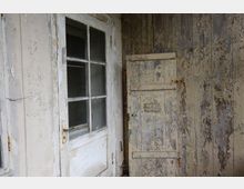 Eine alte, verwitterte Holztür mit Glaseinsätzen neben einer ebenso verwitterten Holzwand, deren Farbe teilweise abgeblättert ist. Die Szene wirkt verlassen und weist sichtbare Spuren von Verfall auf.