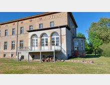 Historisches Gebäude mit einem hellen Anbau, der durch drei große Rundbögen und weiße Fenster geprägt ist. Der Bau steht in einer grünen, parkähnlichen Umgebung, mit Bäumen im Hintergrund und Holzstapeln im Vordergrund.
