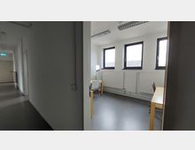 Ein einfacher, heller Büroraum mit drei Fenstern, weißen Wänden, einem grauen Boden und schlichten Holzmöbeln, bestehend aus zwei Tischen und einem Stuhl. Der Raum befindet sich am Ende eines weißen Korridors mit geschlossener Tür.