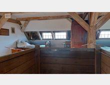 Dachgeschossraum mit Holzfachwerk, eingeschossigen Fenstern und einfacher Einrichtung, darunter ein Metallbett, ein Schrank und ein Tisch. Im Vordergrund befindet sich eine hölzerne Abtrennung mit Absperrkette.