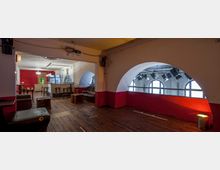 Innenraum eines Veranstaltungsortes mit Holzboden, einer Bar mit Hockern im Hintergrund und einem Bereich mit Sitzmöbeln. Große, gewölbte Fenster mit Bühnenscheinwerfern prägen die helle Rückwand, während Wände teils in Rot und Weiß gestrichen sind.